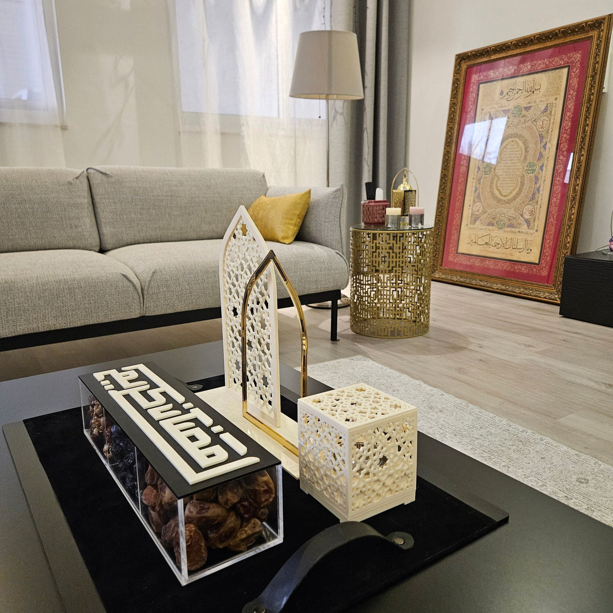 A Ramadan set on a black leather tray: a dates box with a Kufic «رمضان كريم» lid, a masjid lantern and a lattice cube on a living-room table
