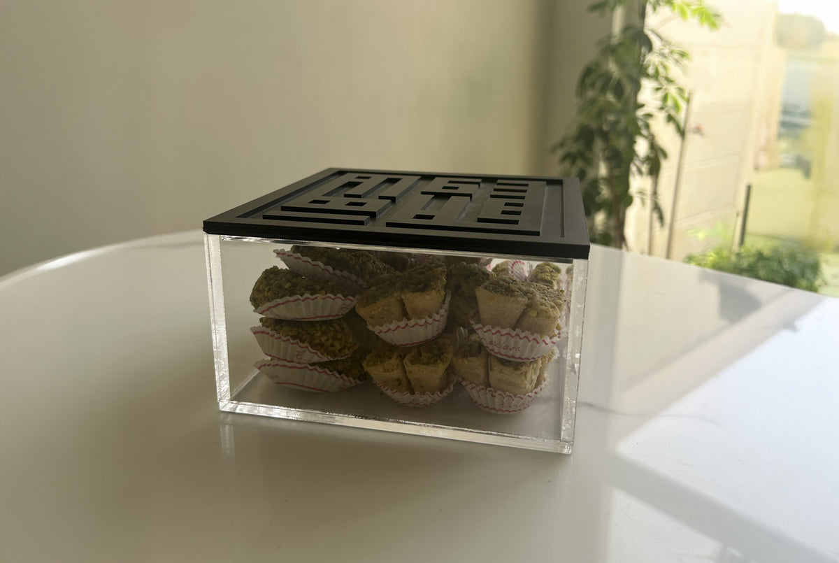 Acrylic box with a black Kufic lid, filled with pastries, on a glass table