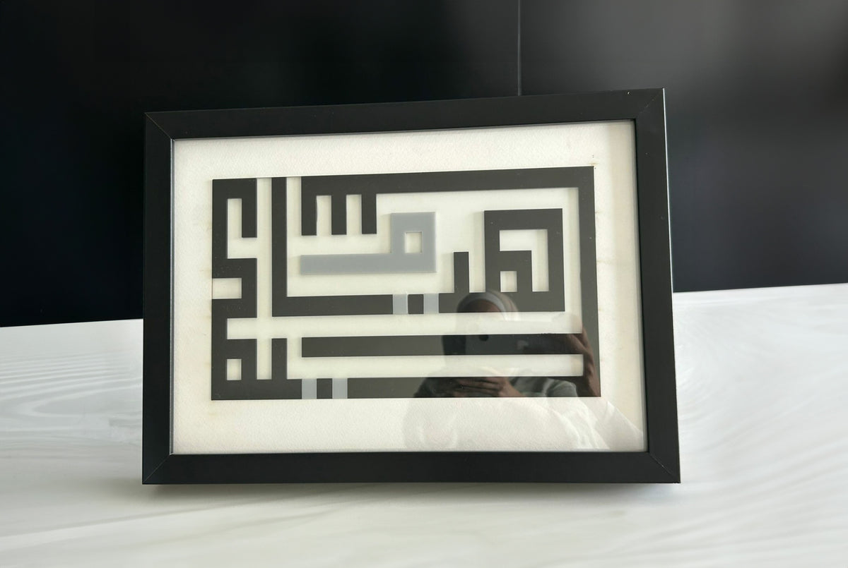 Framed square-Kufic name panel in a black frame, standing on a table