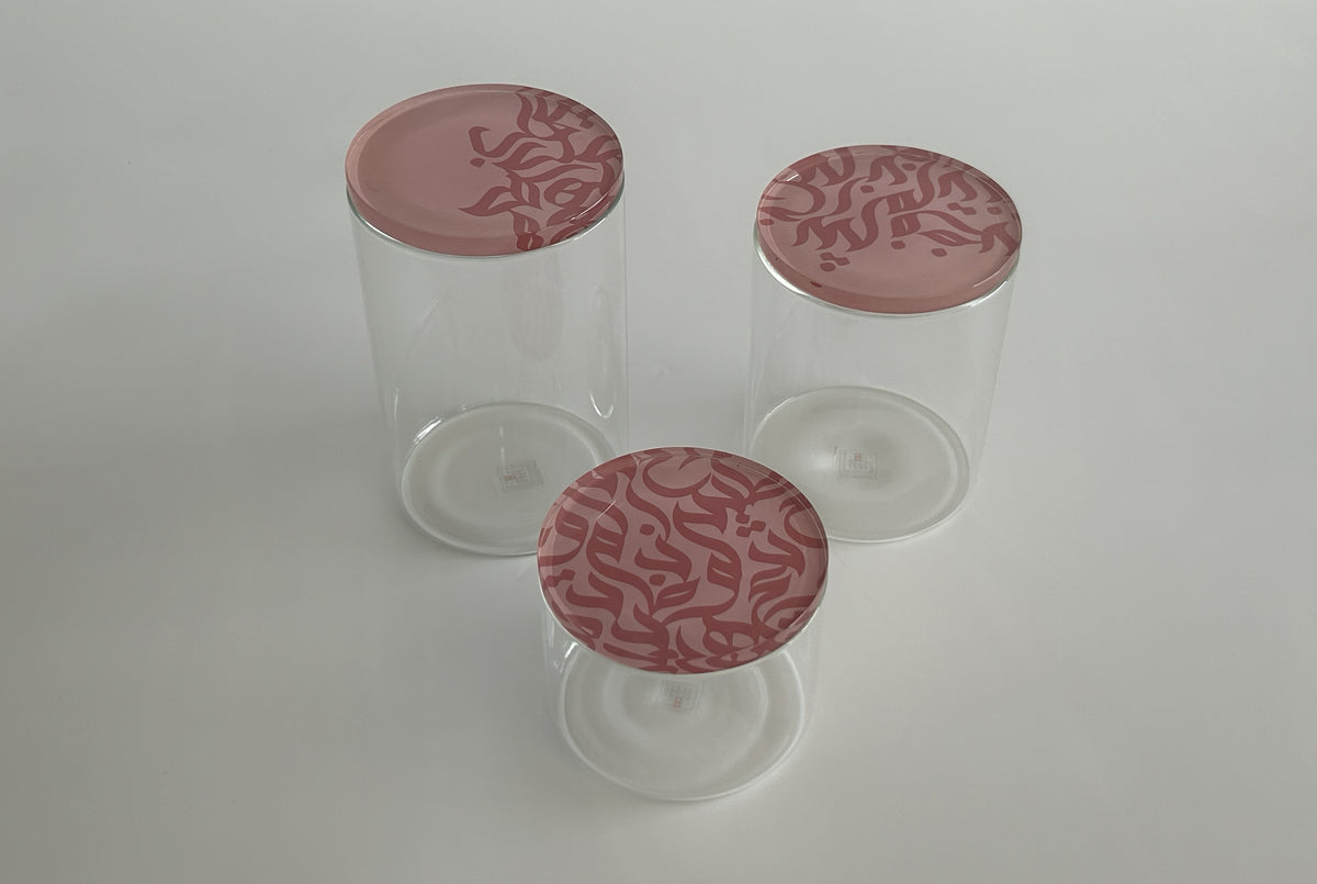Three glass storage jars with pink lids carrying flowing Arabic calligraphy