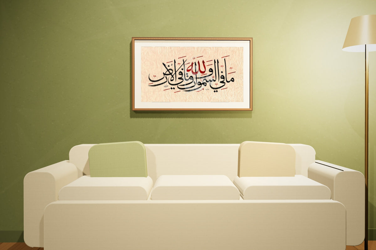 «لله ما في السموات وما في الأرض» بخط أحمد الهواري، 70 × 50 سم، معروضة بمقاسها الحقيقي في إطار جوز داكن في صالة زيتونية اللون — التأطير متاح عند الطلب