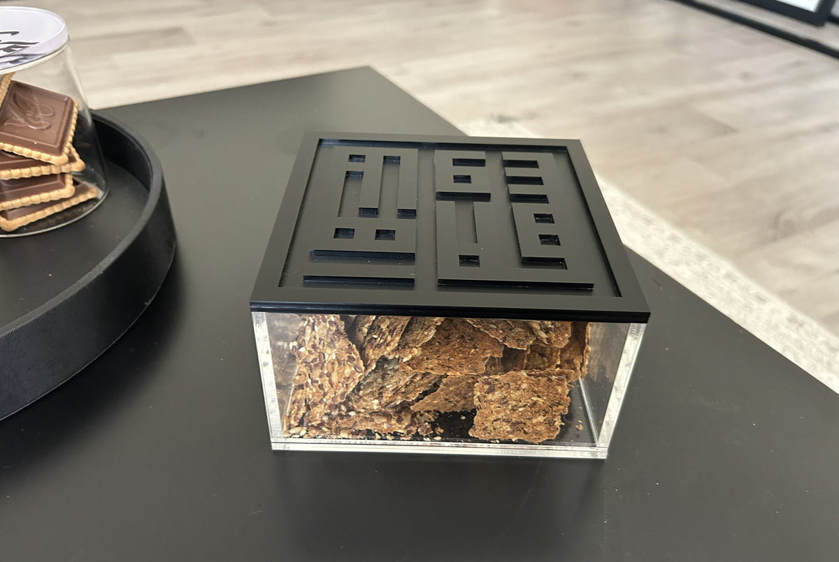 Acrylic box with a black Kufic lid, filled with dates, on a dark table