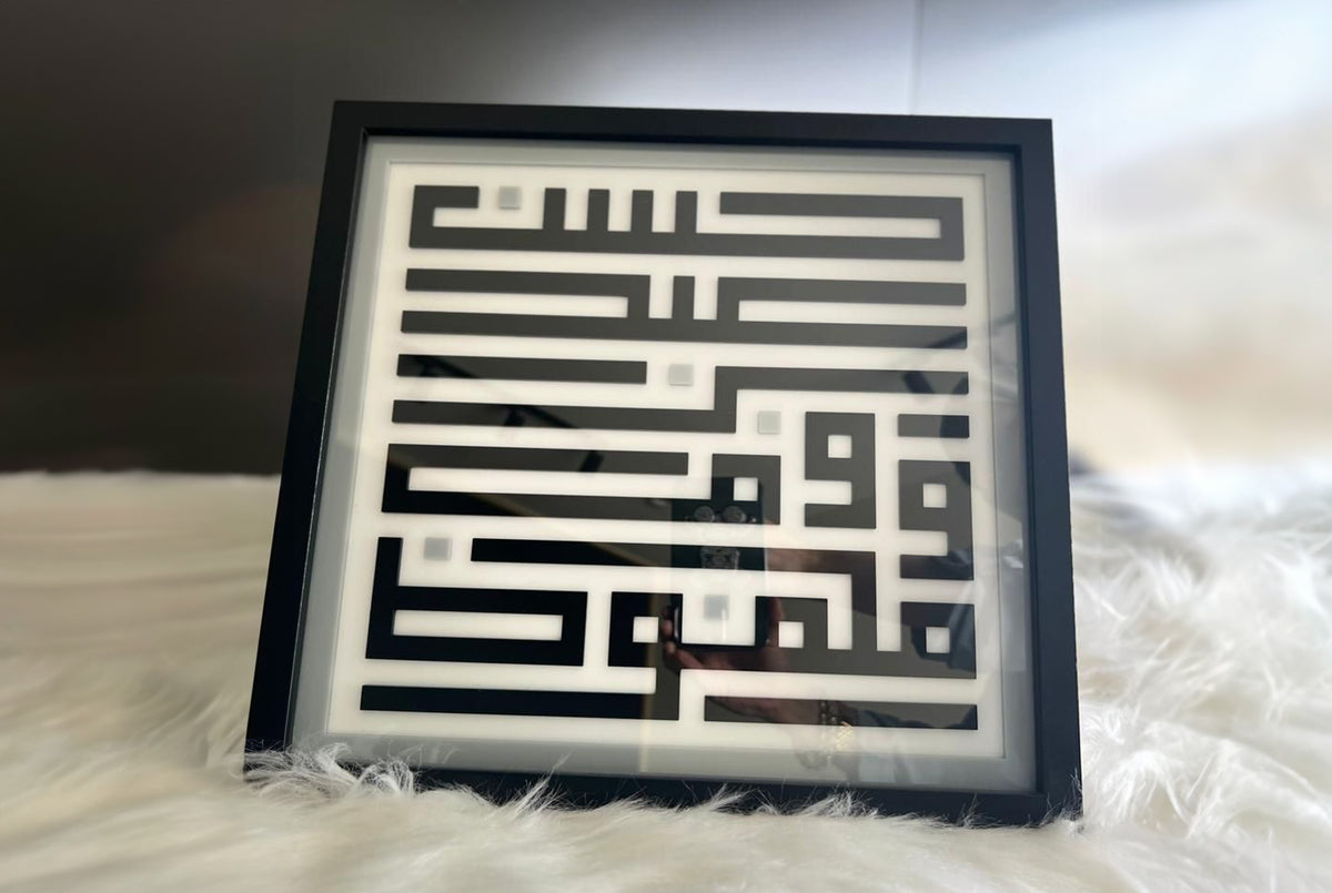Framed square-Kufic name panel photographed at an angle on a soft white rug