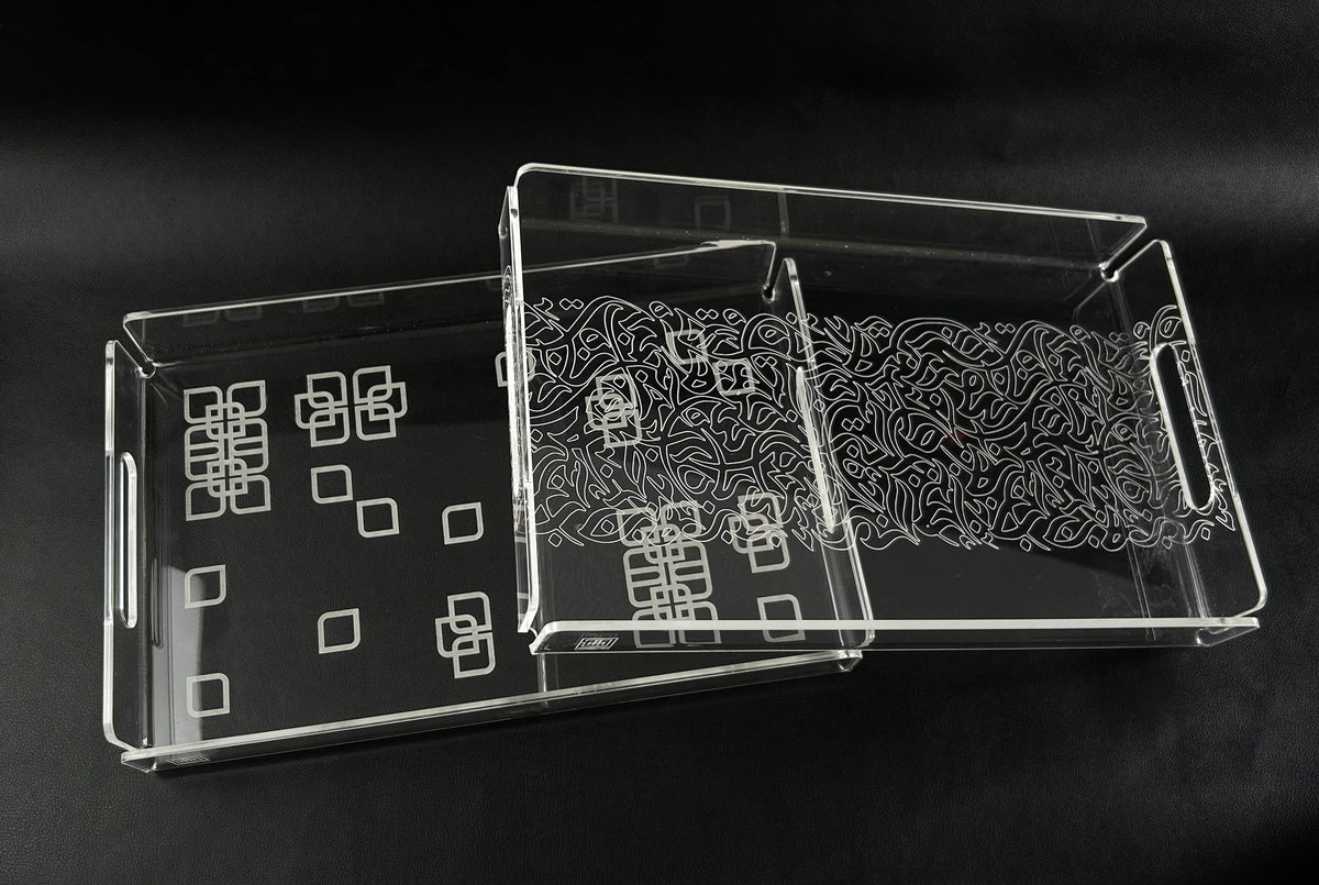 Two clear acrylic trays on a black surface, one with white square motifs and one with dense engraved calligraphy