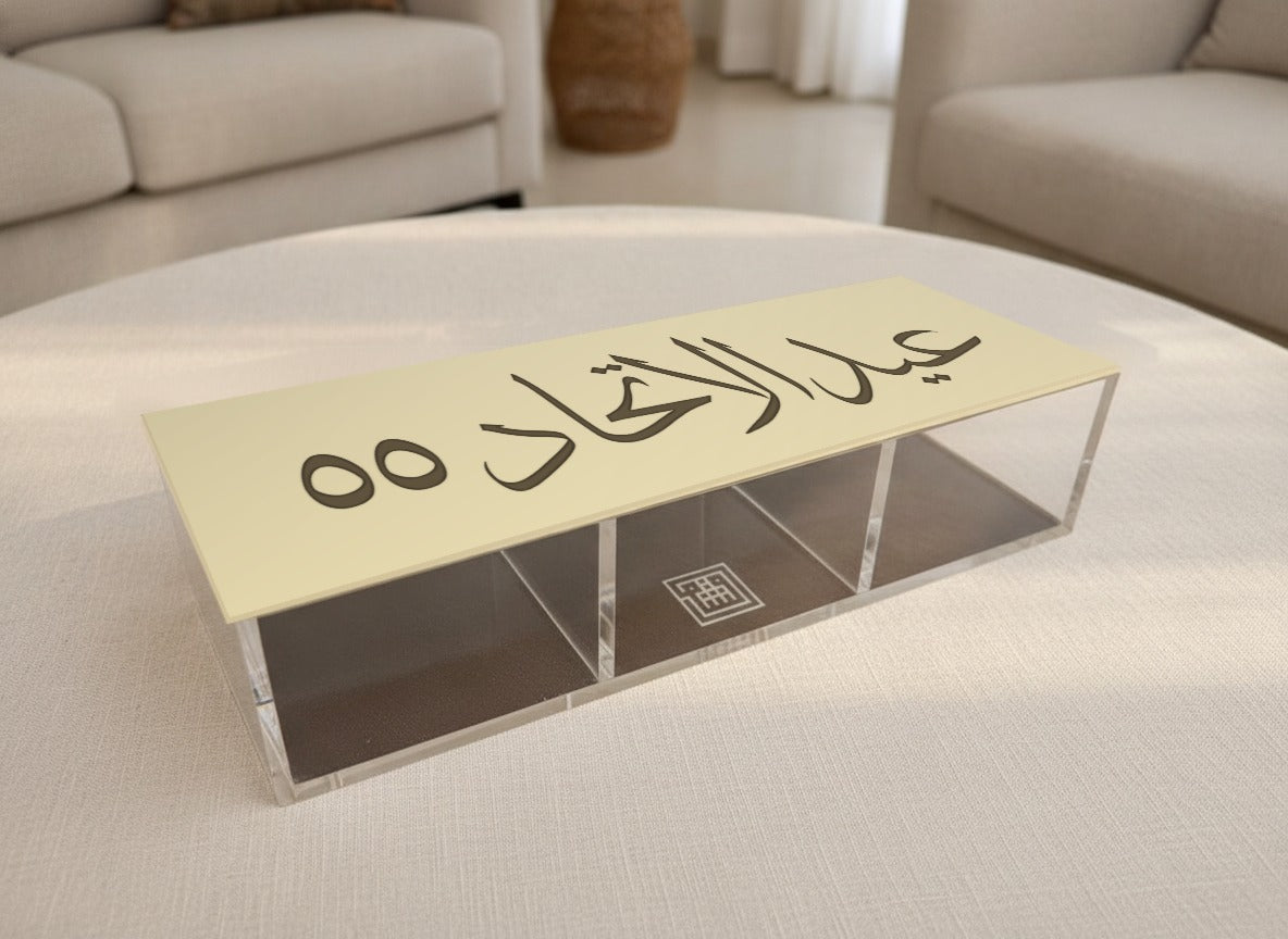 علبة تمر «عيد الاتحاد ٥٥» لليوم الوطني مغلقة على طاولة مجلس مستديرة، العبارة بخط الثلث مفرّغة في غطائها العاجي، وثلاث خانات شفافة تحته